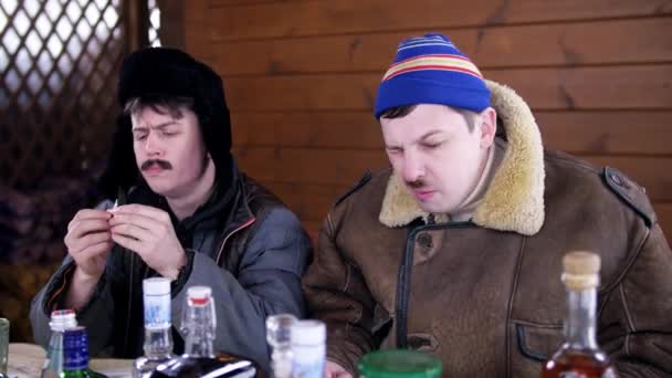 Facet w zimowych ubraniach siedzi z przyjacielem ubrany w kapelusz z klapami wokół stołu — Wideo stockowe