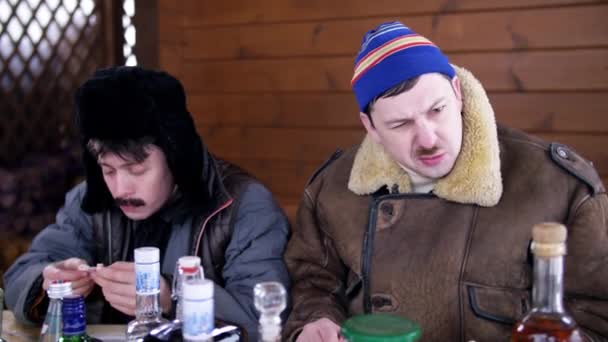 Dois homens amigos em roupas de inverno e chapéus sentar e conversar em torno da mesa cheia de bebidas . — Vídeo de Stock