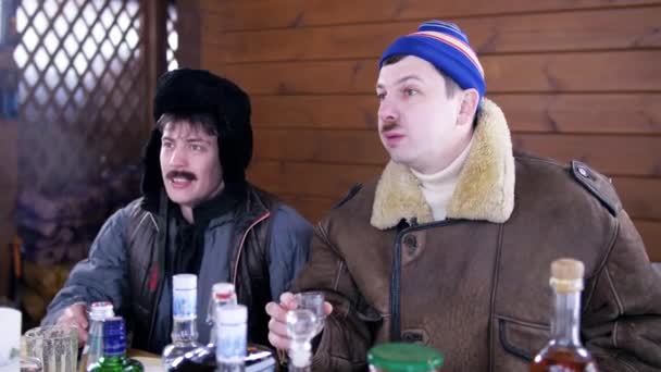 En casa de campo dos hombres fritos en ropa de invierno se sientan alrededor de la mesa y ríen . — Vídeos de Stock