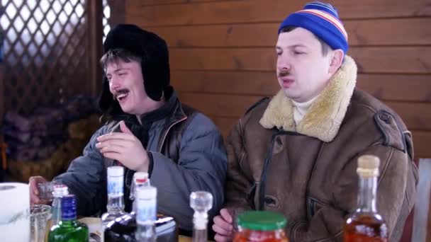 Dois caras vestindo roupas de inverno sentar em torno da mesa, rir e beber álcool . — Vídeo de Stock