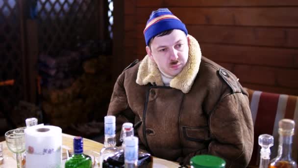 Śmieszne kaukaski facet pijany z wąsy w niebieskim kapeluszu i płaszcz siedzi za stołem — Wideo stockowe
