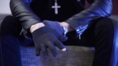 Sıkı deri ceketli adam kanepeye oturur ve yavaş yavaş koyu gri eldiven giyiyor