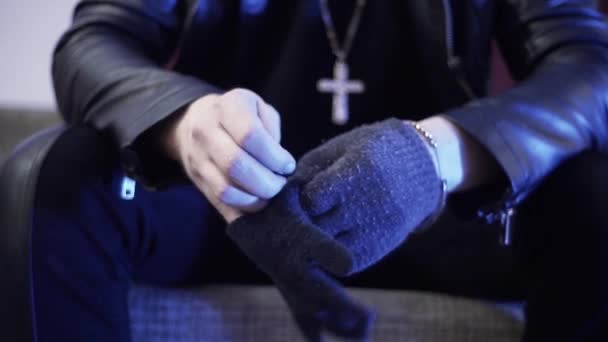 Человек в черной кожаной куртке сидит на диване и медленно одевается в темно-серые перчатки — стоковое видео