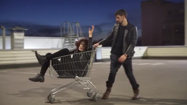 Hombre con chaqueta empuja a otro tipo divertido vestido como mujer en el carrito de la compra . — Vídeo de stock