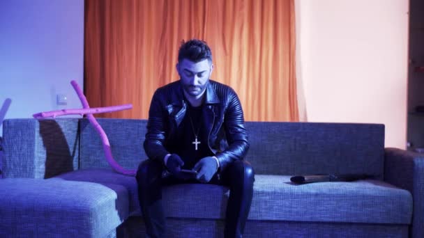 Attractive man with stylish hair cut in leather jacket sits on sofa and sings. — Stock Video