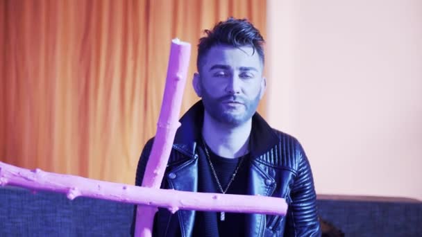 Cute caucasian man in leather jacket sings and dances on sofa holding pink cross — Stock Video