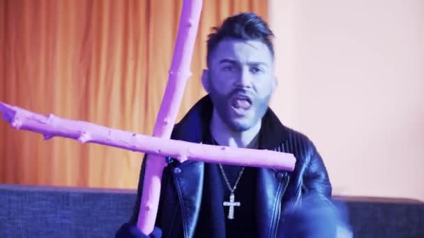 Good-looking male in leather jacket sings and dances on sofa holding pink cross. — Stock Video