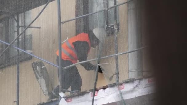SAINT PETERSBURG, RUSSIA - DECEMBER 15, 2018:Construction laborer in orange uniform vest scratches wall with hammer on scaffolding — Stock Video