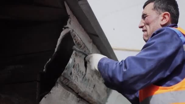 サンクトペテルブルク、ロシア - 4月 10, 2018: オレンジ色のユニフォームベストを着たアジアの男性労働者が汚れた古い木製の壁を壊す. — ストック動画