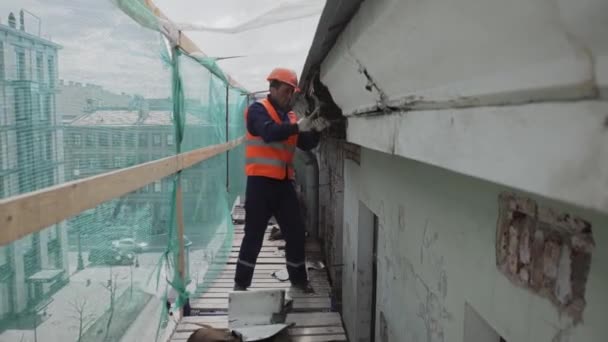 Αγία Πετρούπολη, Ρωσία-10 Απριλίου, 2018: ασιατικό αρσενικό εργαζόμενος σε πορτοκαλί στολή και κράνος σπάει βρώμικο παλιό ξύλινο τοίχο. — Αρχείο Βίντεο