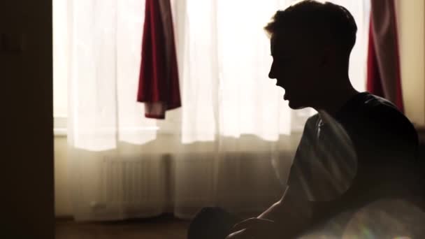 Schöner junger Mann sitzt und singt im Schlafzimmer mit Gegenlicht aus den Fenstern. — Stockvideo