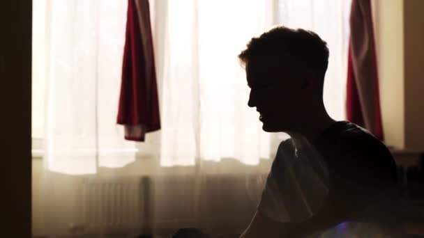 Junger aufgebrachter Mann sitzt und singt im Schlafzimmer mit Hintergrundbeleuchtung aus den Fenstern. — Stockvideo