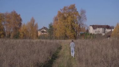 Kamera, kırsal alandaki evlere doğru gri tarlaların arasında yürüyen adamı gösteriyor..