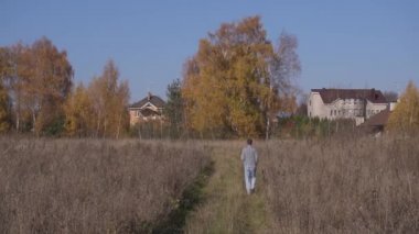 Kamera, yetişkin bir adamın kırsal alandaki evlere doğru gri tarlaların arasında yürüdüğünü gösteriyor..