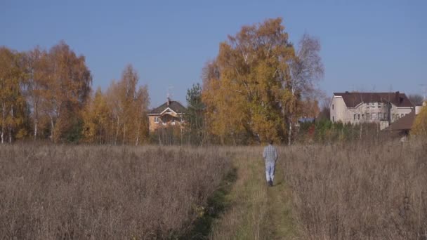 Mann läuft langsam zwischen grauen Feldern auf große Häuser im ländlichen Raum zu. — Stockvideo