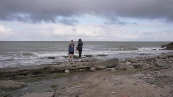 Two women stand at coastline with waves falling on shores on windy summer day. — Stock Video