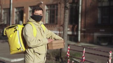 Siyah maskeli, ceketli, sarı sırt çantalı, pizza kutulu beyaz erkek teslimatçı güneşli bir havada sokakta kameraya bakıyor..