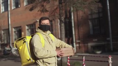 Siyah maskeli, ceketli, sarı sırt çantalı, pizza kutulu beyaz erkek teslimatçı güneşli bir havada sokakta kameraya bakıyor..