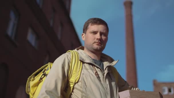 黄色のバックパックと天気の良い日に通りに立って カメラを見ているピザボックスを持つジャケットの白人配達人 — ストック動画