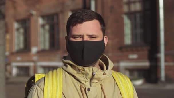黒いマスクの白人男性の配達人とカメラに黄色のバックパックを持つジャケットは 彼のマスクを脱ぎ 深呼吸を取る — ストック動画