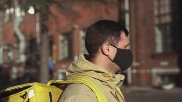 黒いマスクの白人男性の配達人とカメラに黄色のバックパックを持つジャケットは 彼のマスクを脱ぎ 深呼吸を取る — ストック動画