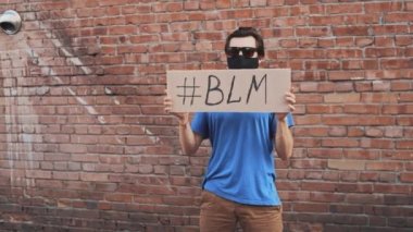 Maskeli beyaz adam kırmızı duvara karşı duruyor elinde karton afiş ve üzerinde de BLM etiketi var. Tek protesto.