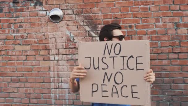 マスク中の白人男性は 碑文と手に段ボールのポスターと赤い壁に対して立っている 正義の平和はありません 抗議1件 — ストック動画