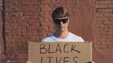 Güneş gözlüklü ve maskeli beyaz adam elinde karton bir posterle kırmızı duvara karşı duruyor. Siyah Yaşamın Önemi. Tek protesto.