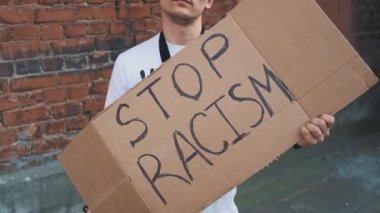 Maskeli beyaz adam elinde karton posterle kırmızı duvara karşı duruyor. Üzerinde 