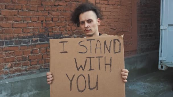仮面の白人男性は赤い壁の反対側に立っており 手には段ボールのポスターが刻印されています 私はあなたと一緒に立つ 抗議1件 — ストック動画