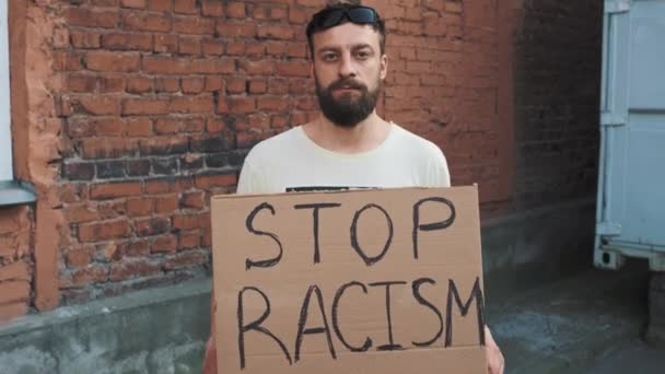 戴面具的戴胡子的高加索人站在红墙上 手上拿着写有 停止种族主义 字样的纸板海报 一次抗议 — 图库视频影像