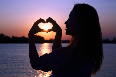 Kız ellerini, batan güneşe karşı kalbin işaretini gösterir.
