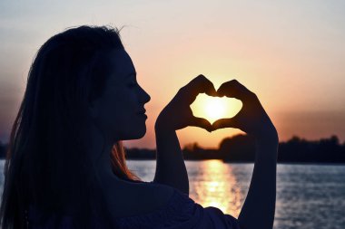 Kız ellerini, batan güneşe karşı kalbin işaretini gösterir.