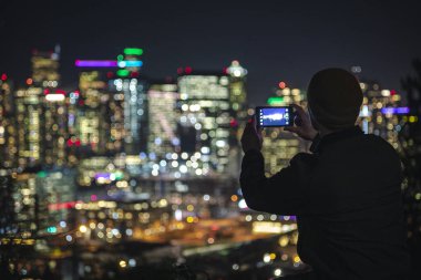Sosyal medya için fotoğraf çekerken cep telefonu tutan adam ile gece kentsel binalardan renkli bulanık arka plan
