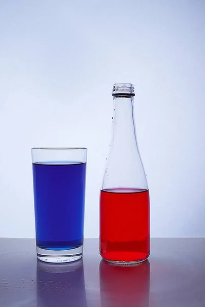 Fles en glas. waarin de rode en blauwe vloeistoffen op een witte ondergrond worden gegoten — Stockfoto