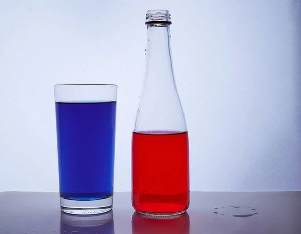 Fles en glas. waarin de rode en blauwe vloeistoffen op een witte ondergrond worden gegoten — Stockfoto