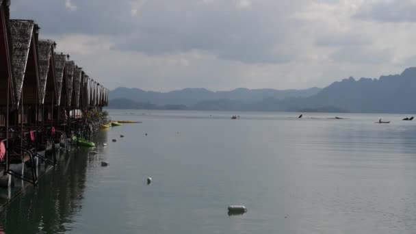 Verschillende bungalows staan op een meer met rieten daken. Mensen zwemmen in een kano. Kajaks zijn geparkeerd op de pier. Lake Cheow LAN. Khao Sok Thailand. — Stockvideo