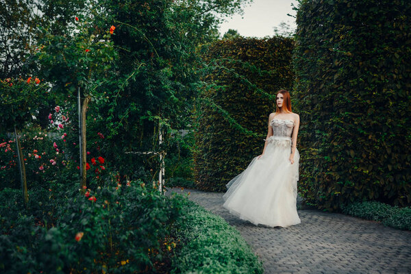 Young beautiful model is posing in a long ivory dress in the garden with a crown on her head