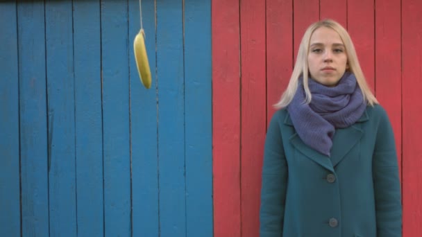 カラフルな木製エコ背景の女の子 女の子ピンク木製エコ背景の上に立つし カメラに見える文字列にバナナが上から来るし 女の子が指でバナナをプッシュします 拒絶反応 — ストック動画