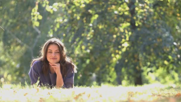 公園で女の子 柏木は草の上に横たわって そっとカメラに探しています 彼女は 彼女の笑顔はかわいいし 愛情のある夢を見る 背景には木が 肯定的な気分 — ストック動画