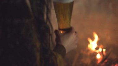 Ateşin yanında bir bardak bira. Kız elinde bira bardağını şenlik ateşinde tutar ve içer. Geceleri arka bahçede ateşle rahatlayın. Alkollü içecek.