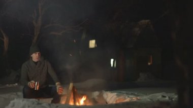 Ateşin yanında adam. Adam ateşin yanındaki metal bardaktan sıcak içecek içer. Duman ve kıvılcımlar birbirinden ayrı uçar. O sıcak giysiler giymiş. Arka bahçede geceleri rahatlayın. House arka planda duruyor. Kış.