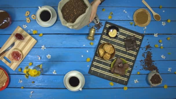 女性の手はテーブルの上に焙煎コーヒー豆の袋を置きます スローモーション トップビュー テーブルの上に別のスナックがあります ケーキ ビスケット ジャム チョコレート製品と甘いチェリーとパン カメラが右から左に移動する — ストック動画