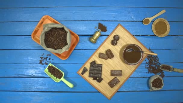 蓝色木桌上的咖啡和巧克力甜点 顶部视图 桌子上是烤咖啡豆在袋子里 研磨咖啡在碗里 速溶咖啡在碟子 桌子上还有不同的巧克力产品 — 图库视频影像