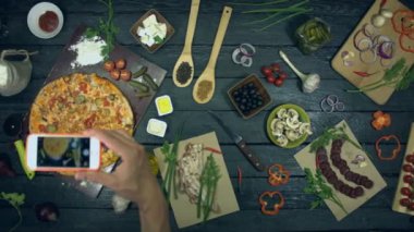 Ekolojik siyah arka planda çizburger pizza. Adam telefonla masaya pizza fotoğrafı çeksin. Pizza malzemelerden oluşur: salatalık, baharat, domates, et, tavuk, sos, peynir.