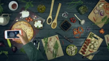 Ekolojik siyah arka planda peynirli pizza. Kız telefonla peynirli pizza nın fotoğrafını çeker. Pizza malzemelerden oluşur: farklı sheese, baharat ve domates. Yemek pişirmek ve pizza yemek için masada birçok ürün vardır.