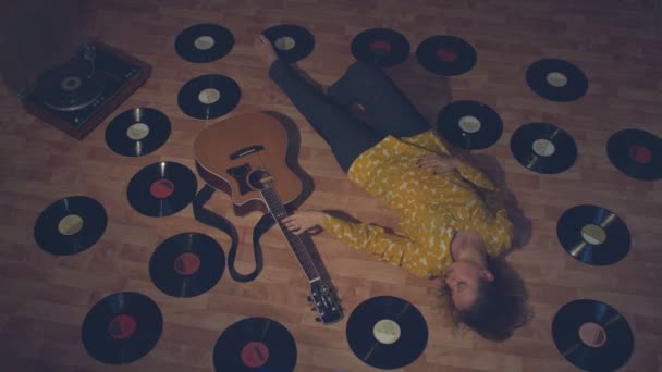 レコードの中でギターを持つ女の子 その少女は床に横たわっている 彼女の隣には 古いレコードとターンテーブルがたくさんあります 彼女はアコースティックギターの弦に触れる — ストック動画