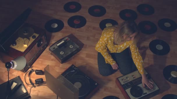 音を再生し 録音するためのヴィンテージデバイスに囲まれた女の子 その少女は床に座っている 彼女の隣には 60年代 70年代 80年代のボビンテープレコーダー 古いターンテーブルとレコードがあります 彼女はテープレコーダーでテープをまっすぐにし — ストック動画