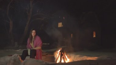 Kız oturur ve kameraya gülümser. Kız oturur, gülümser ve ateşe bakar. Kalın giysiler giydi..