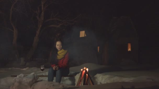 女孩一个人坐在篝火边的院子里 晚上一个人喝酒 背景是房子 — 图库视频影像